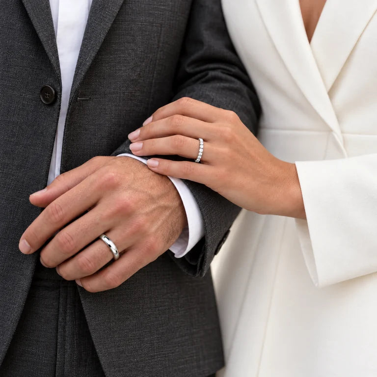 A standing couple in tailored charcoal and ivory clothing, showing a man’s polished white-gold wedding band and a woman’s diamond eternity band