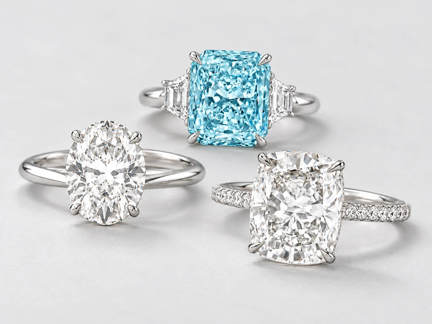 Oval diamond solitaire and elongated cushion diamond ring with a radiant-cut aquamarine three-stone platinum ring behind them on light gray