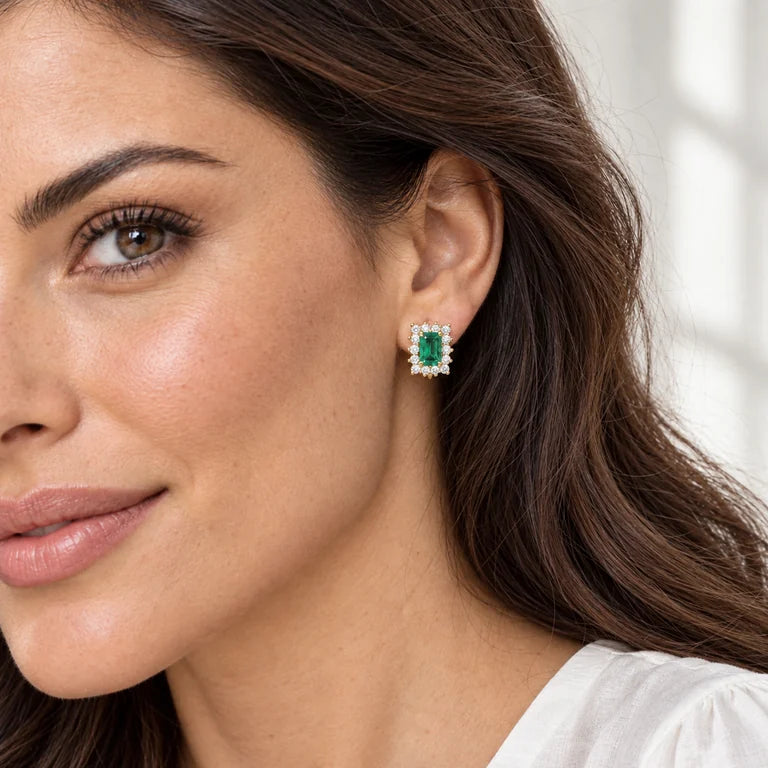 Model wearing emerald-cut green emerald and diamond halo earrings in yellow gold