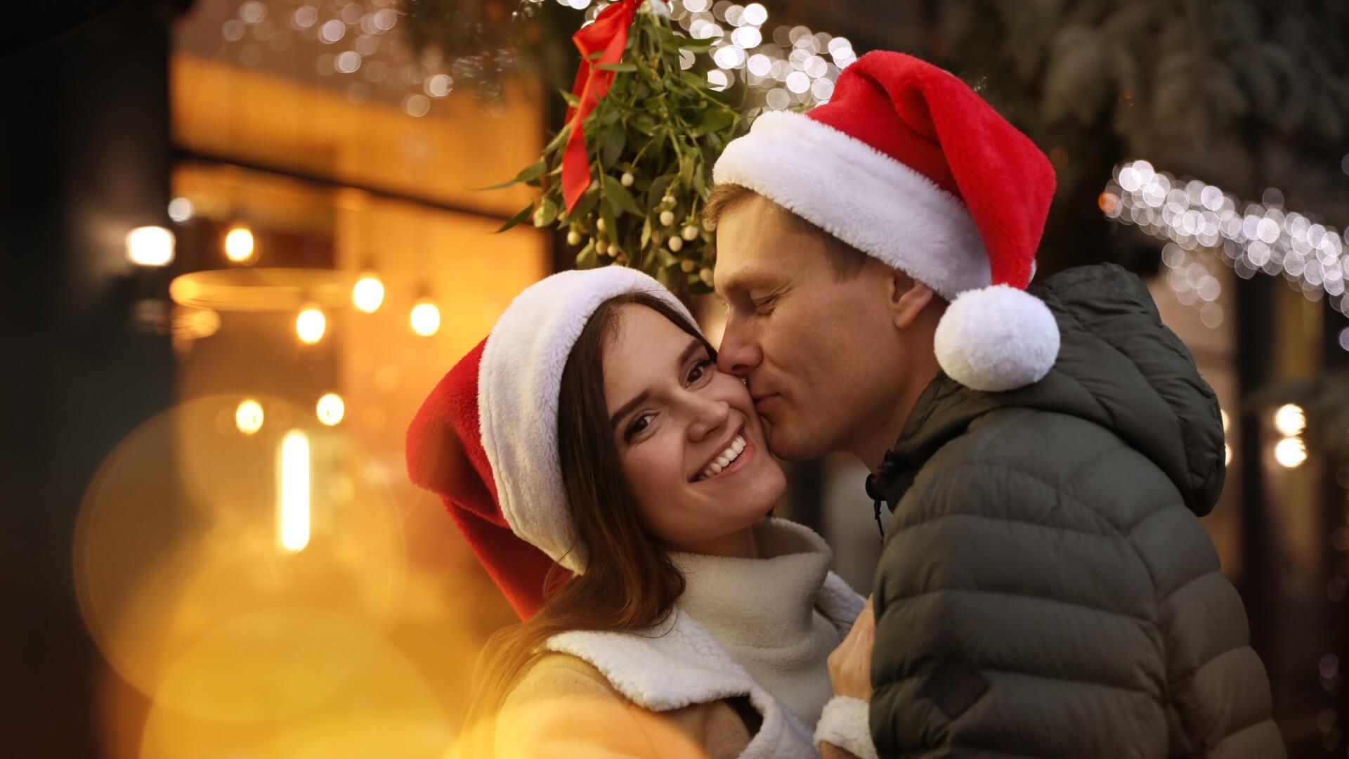 a couple wears Santa Claus hats as they kiss under the mistletoe