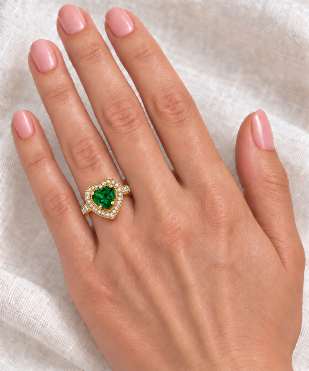 10x10mm heart-shaped lab emerald and white diamond halo ring in 14k yellow gold, 3.66ct — correctly scaled at base of left ring finger on ivory linen