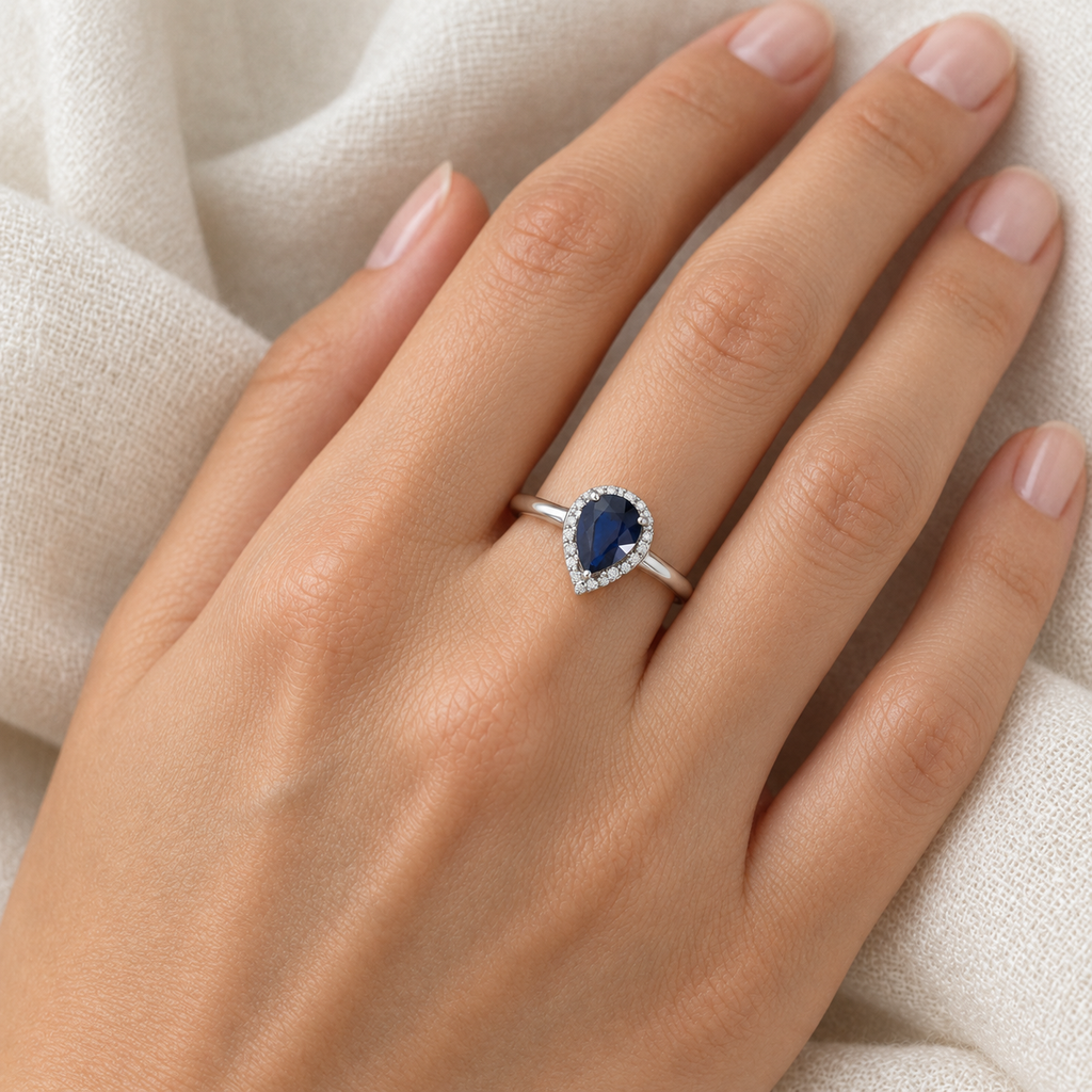 Natural pear blue sapphire and diamond halo ring in 14K white gold on left ring finger, ivory linen closeup