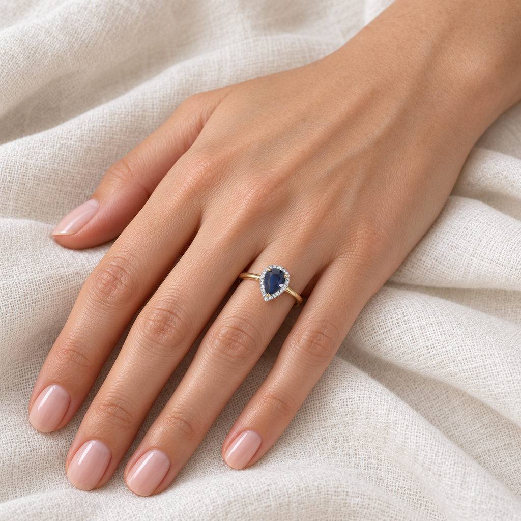 Natural pear blue sapphire and diamond halo ring in 14K yellow gold on left ring finger, ivory linen closeup