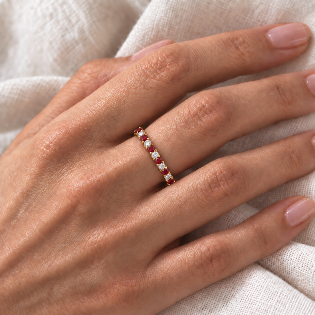 Lab-grown red garnet and lab-grown diamond eternity ring in 14k yellow gold, correctly scaled 2.5 mm alternating band, worn at the base of the left ring finger on ivory linen