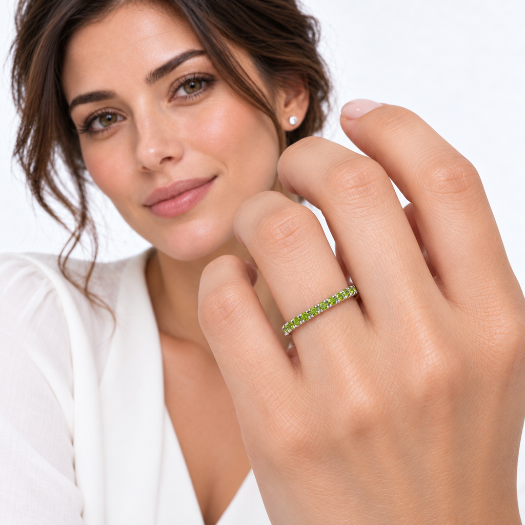 Face-visible brunette model in ivory blouse with foreground hand wearing fine 2 mm half-eternity ring with 19 round faceted natural peridots, 0.65ct, high-polished 14k white gold.